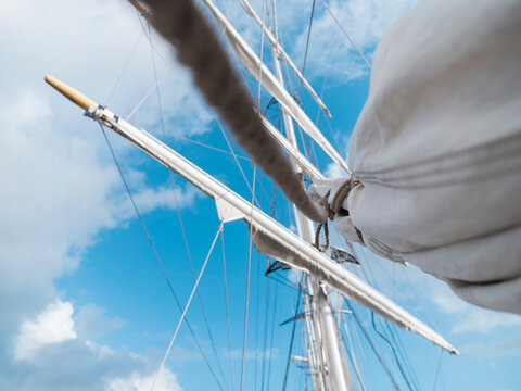 A tilted view into the rigging of a tall ship, yard and furled sail sweeping past the mast and a bright sky full of clouds.