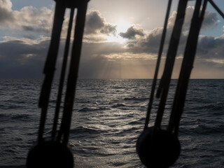 The sun stands behind cloud above a wide sea, framed by the blurred dark shapes of a ship's rigging in the foreground. © Victor