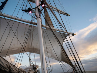 Square sails stand full of wind above the deck of a tall ship at dusk, pink stage lights glowing on the mast against pastel clouds. © Victor