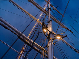 The lit mast of a sailing ship stands against a dark blue night sky, warm floodlights catching the furled sails and rigging. © Victor