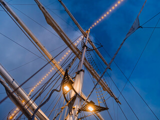 Two strings of warm bulbs run diagonally through the rigging of a sailing ship below floodlights and a deep blue sky. © Victor