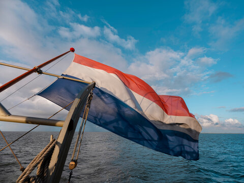 The Dutch flag flies from the stern of a sailing ship above bright blue water, with a wooden spar and block alongside.