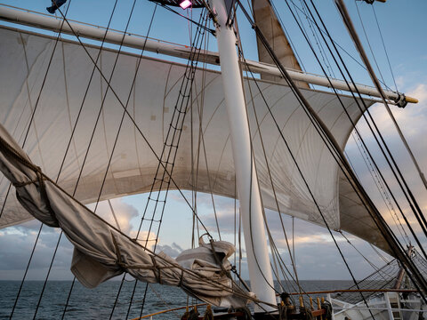 Square sails fill with wind above a furled sail and the sea beyond, seen from the deck of a tall ship in evening light.