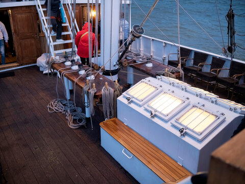 Guests gather on the lit deck of a sailing ship at blue hour, wicker chairs, glowing skylights and coiled ropes on the belaying rail.
