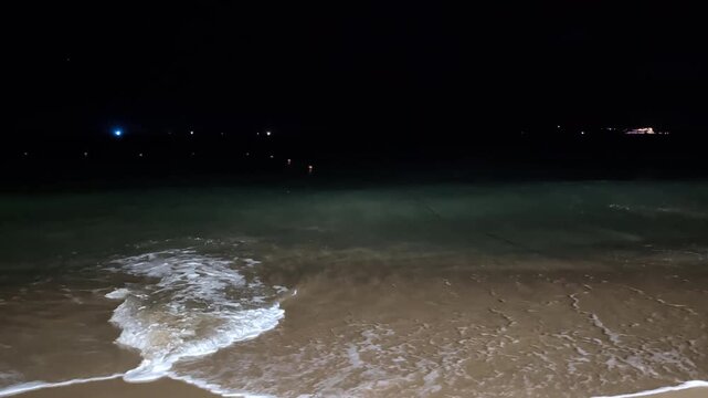 Night shot of a sandy beach as sea waves roll in. The surging wave stirs up sand from the bottom, while colliding undercurrents and incoming waves crash into each other, creating thick white foam.