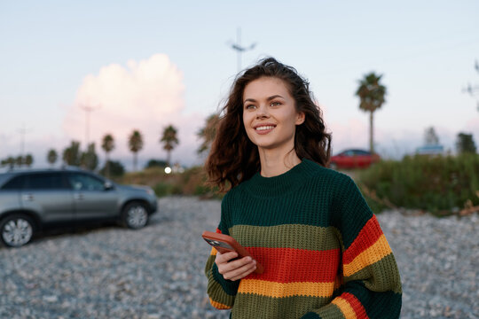 A smiling young woman in a striped sweater holds a smartphone outdoors. A car is visible in the background of the street. Concept for modern lifestyle