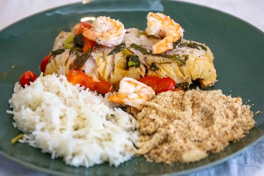 Moqueca capixaba, traditional Brazilian seafood stew with snook fish, shrimp, tomato and coriander, served on a green plate with white rice and farofa.