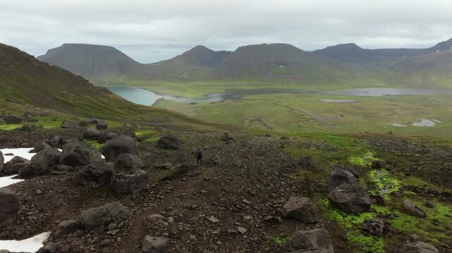 Aerial drone push in shot of a lone hiker traversing a path in the vast, remote Hornstrandir nature reserve on Iceland's Westfjords, surrounded by lush green valleys and majestic table mountains.