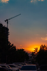 A high-rise building under construction with a towering construction crane silhouetted against a dramatic vibrant orange and blue sunset sky above a busy city street. © Dina Luk
