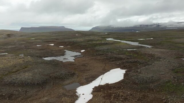Aerial drone footage pushes in on a lone hiker trekking across a remote, rugged landscape in Hornstrandir, Iceland. Features mountains, streams, and snow patches under an overcast sky.