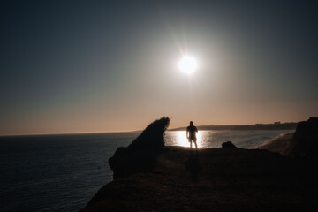 Person stands on cliff by the ocean during sunset hours with bright sun above © Bohdan