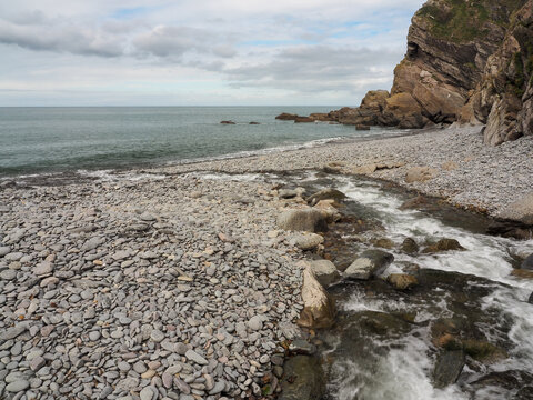 The River Heddon flows across a pebble beach to meet the sea beneath rugged coastal cliffs at Heddons Mouth on the Exmoor coastline in Devon, UK