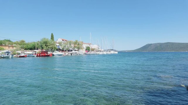 The Steni Vala harbor in Alonnisos, Greece, showing clear blue Aegean Sea water, moored sailboats and fishing boats, traditional waterfront houses, and a rocky coastline