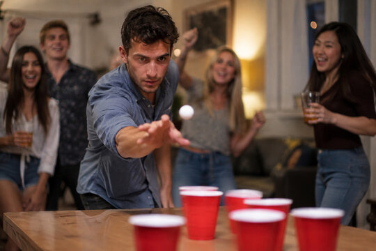 Young man reaches toward a ping pong ball during a lively cup-toss party game as friends cheer behind him in a warmly lit living room.