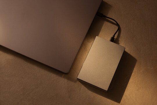 External hard drive connected to a laptop computer on a beige surface with warm soft lighting, showing modern data storage technology, file backup process, digital privacy, and workspace setup.
