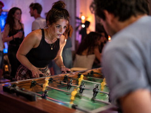 Focused young woman grips the rods of a tabletop soccer game while facing an opponent at a lively indoor party with blurred guests behind.
