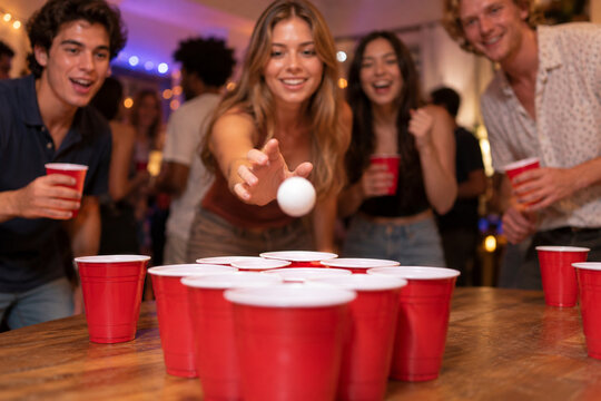 Low table-level view of red cups arranged for beer pong as a young woman takes her shot and friends react around the game table.