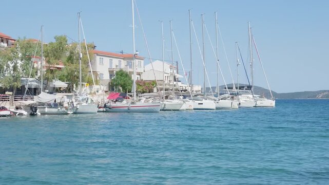 A tranquil scene in Steni Vala harbor on Alonnisos island, Greece, showing sailboats and yachts docked along the waterfront with clear blue water and coastal village buildings