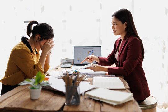 Stressed young employee covering face with hands while receiving an angry reprimand from a demanding female boss during a difficult meeting in a busy office environment
