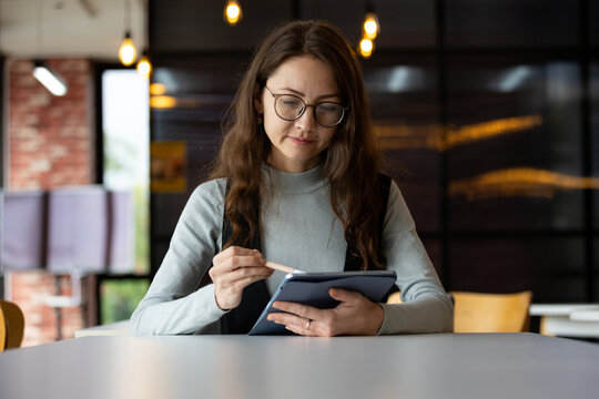 Focused woman with glasses using tablet and stylus at modern workspace, showing digital work, technology, productivity, learning and professional concentration indoors
