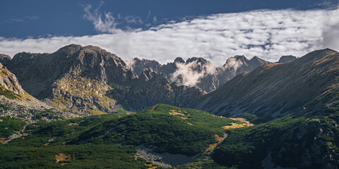 Panoramic view of the High Tatras featuring rugged mountain ridges, alpine terrain and dwarf pine vegetation under dramatic clouds and clear blue sky in Slovakia. © ukasz