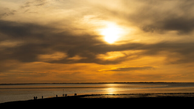 Dramatic sunset silhouette scene along the Wadden Sea shoreline, showing a wooden breakwater structure extending into the golden reflective water with people enjoying the view.