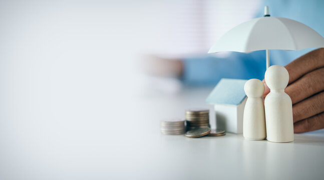 Hand holding a white umbrella protecting wooden family figures, house model, and coin stacks on desk with copy space, concept of insurance, family protection, security, and financial planning.