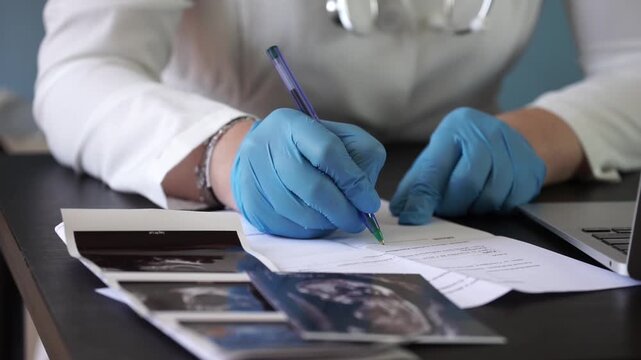doctor in blue gloves writing on document with ultrasound photos on desk in medical office closeup, concept of obstetrician, medical consultation, pregnancy checkup