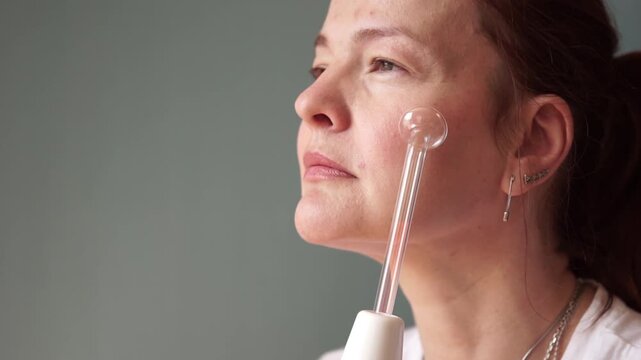 woman using darsonval electric device on cheek gazing away in bright studio closeup, concept of skincare routine, cosmetology, aesthetic medicine. High quality photo