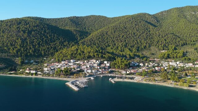Aerial view of Neo Klima, a coastal village on the island of Skopelos, Greece. The marina with docked sailboats, the shoreline, and the village nestled at the base of lush green hills