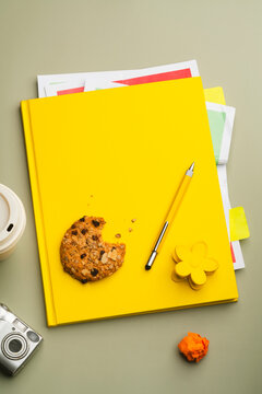 Messy table top with yellow book, pencil and a bitten cookie.