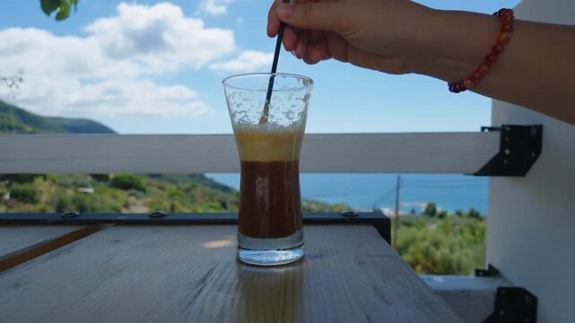 A hand stirs a glass of iced coffee on a wooden table, set on an outdoor terrace overlooking lush green hills and the blue Aegean Sea in Glossa, Skopelos, Greece