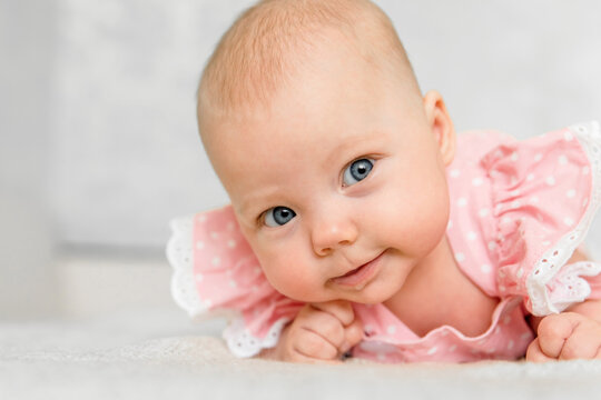 Baby girl 5-6 months old in pink clothes lies on tummy at home, smiles and looks at camera, cheerful portrait with blue eyes