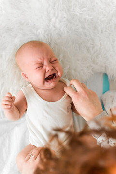 Baby 5-6 months old cries while mother puts on a white bodysuit at home, authentic everyday baby care