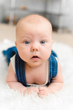 Baby 5-6 months old in blue overalls lies on tummy on a white blanket at home, exercising muscles and looking at camera