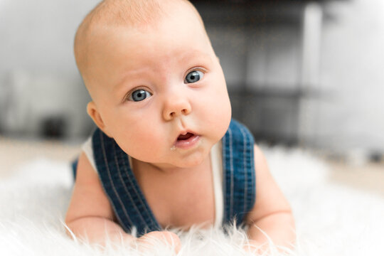 Baby 5-6 months old in blue overalls lies on tummy on a white blanket and looks at camera in a close home portrait