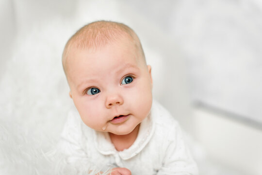 Baby 5-6 months old in white clothes lies on tummy at home and looks at camera, bright portrait with blue eyes
