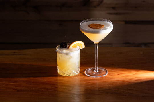 Beautiful Whiskey Sour served two ways, up and on the rocks with a warm wooden surface and background texture.