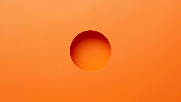 A perfectly circular hole cut into a vibrant orange paper surface, creating a deep shadow and a three dimensional effect on the flat background