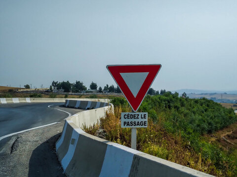 Red triangular yield sign standing beside a curved asphalt road with concrete barriers under bright clear sky on a sunny day in the Moroccan countryside