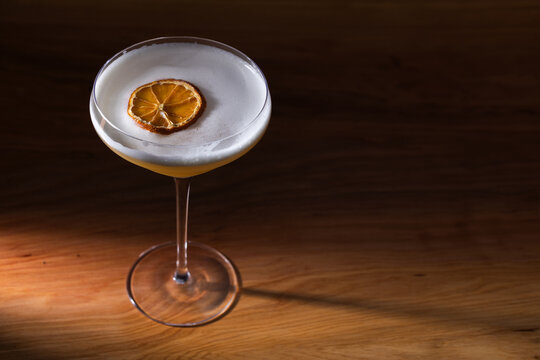 Beautiful Whiskey sour served up in a couple glass garnished with a dehydrated orange wheel with a warm wooden surface and background texture.