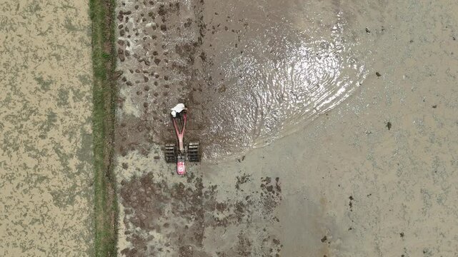 An aerial view of a Asian farmer operating a hand tractor to plow a muddy rice field prepare the soil for agricultural planting under bright natural sunlight in a rural traditional countryside.
