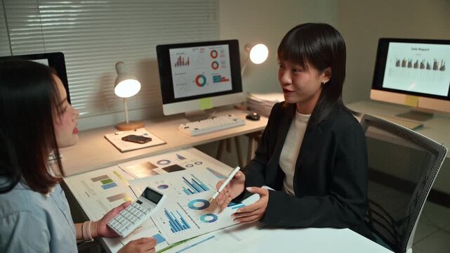Two dedicated young Asian businesswomen collaborating on a project, analyzing financial data and charts on documents and computers while working overtime together in a modern office at night