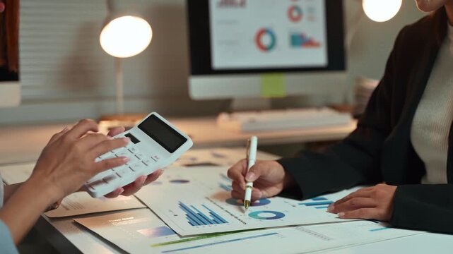 Two businesswomen collaborating on a financial project, analyzing charts and graphs on paper while using a calculator to check the company's performance and prepare a detailed report