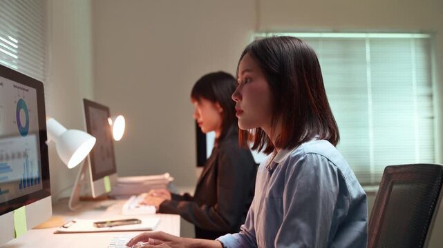 Two focused Asian women colleagues working late at night in the office, typing on keyboards and analyzing financial charts and graphs on their computer monitors to meet a project deadline