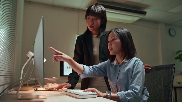 Two young Asian businesswomen working late at night in the office, with a manager assisting and training her new employee on a computer, demonstrating teamwork and professional collaboration