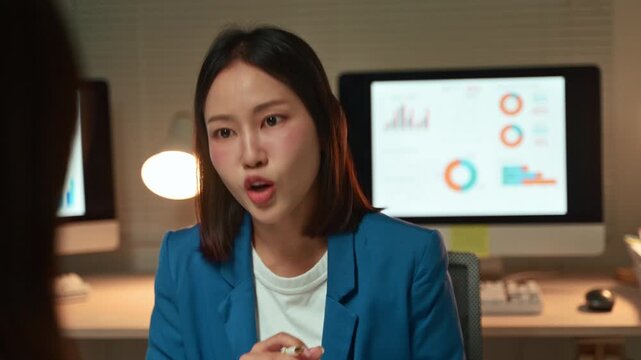 Businesswoman seriously discussing ideas with her colleague, gesturing during a meeting in a modern office at night, while working late to meet a project deadline with computers in the background
