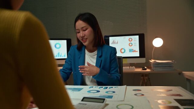 Two young Asian businesswomen meeting, talking, and discussing their company's financial data and marketing plans on a paperwork graph and chart in a conference room at the office late at night