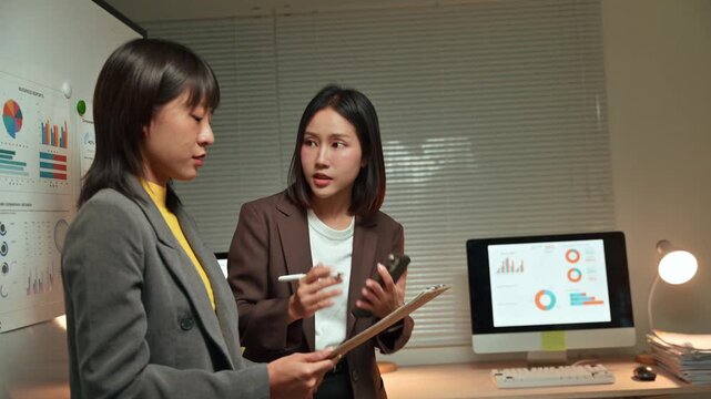 Two dedicated Asian businesswomen working late at night, collaborating on a project by analyzing financial data on a clipboard and computer screen in a modern office environment