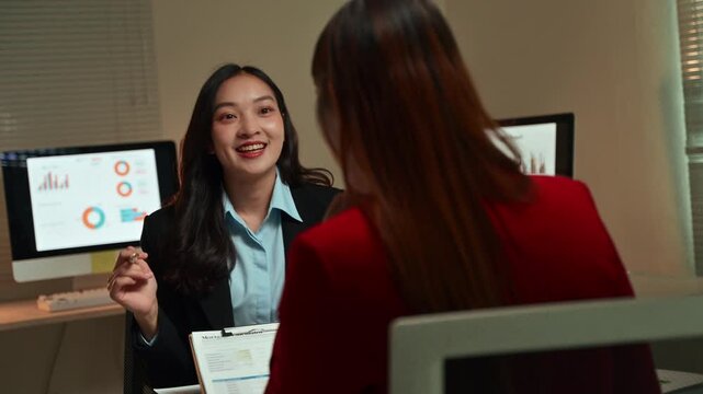 Two young asian businesswomen discussing financial data on a clipboard during a late-night meeting in a modern office, collaborating on a project and analyzing graphs for a successful strategy